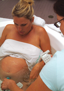 Heavily pregnant woman sitting in a large bath while a midwife presses a stethoscope to her abdomen to monitor the baby.